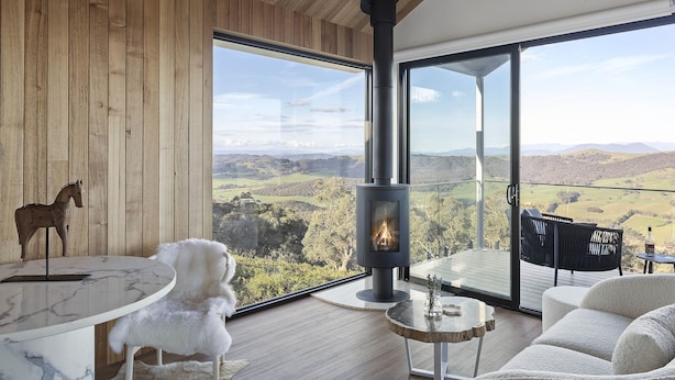 Una habitación moderna con un ventanal grande, chimenea, una mesa redonda y un sillón blanco mullido.