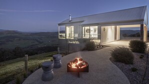 Una casa moderna con un brasero en primer plano, un gran ventanal que da a una terraza y vistas al campo.