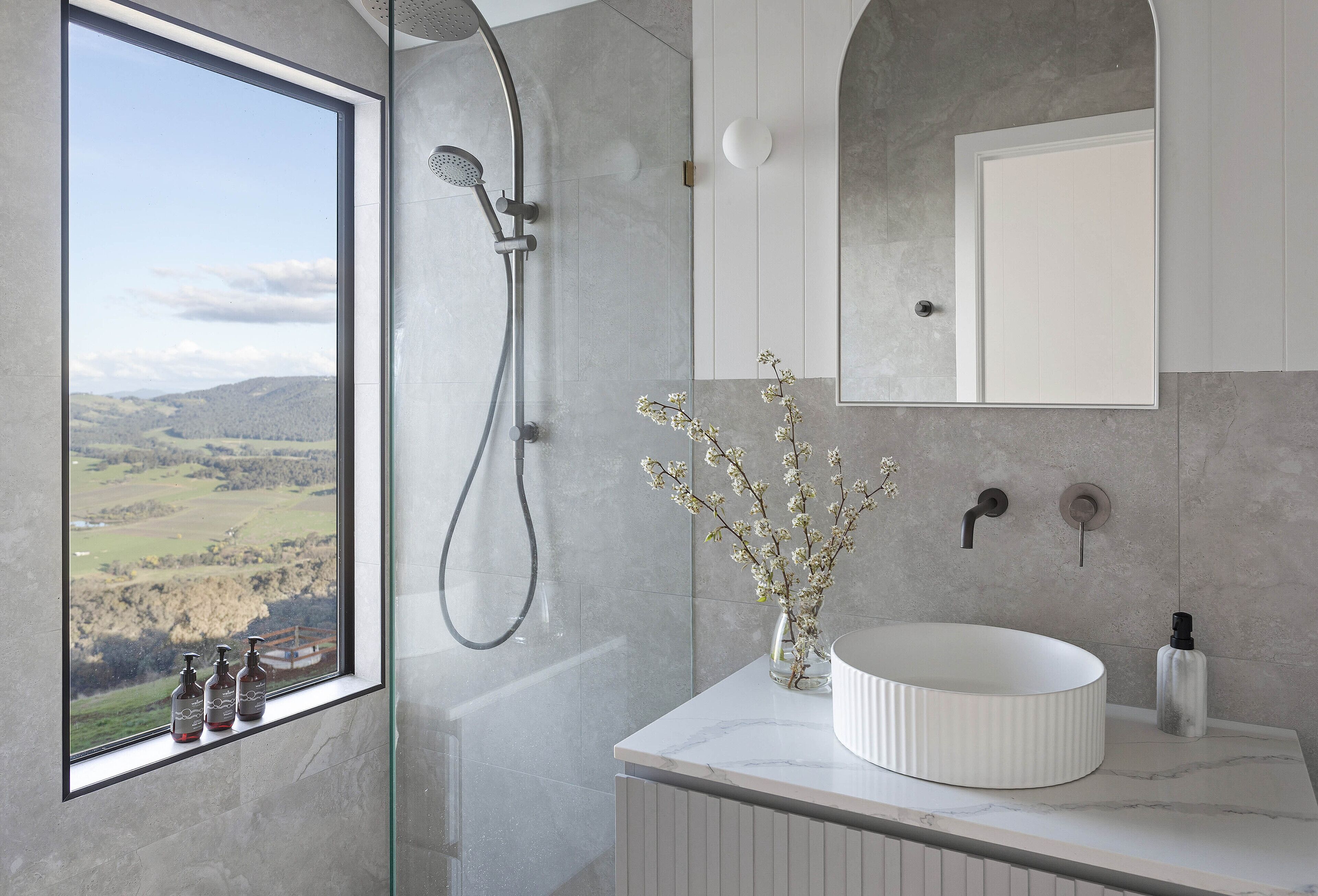 A bathroom with a large window, a shower, a sink, and a mirror.