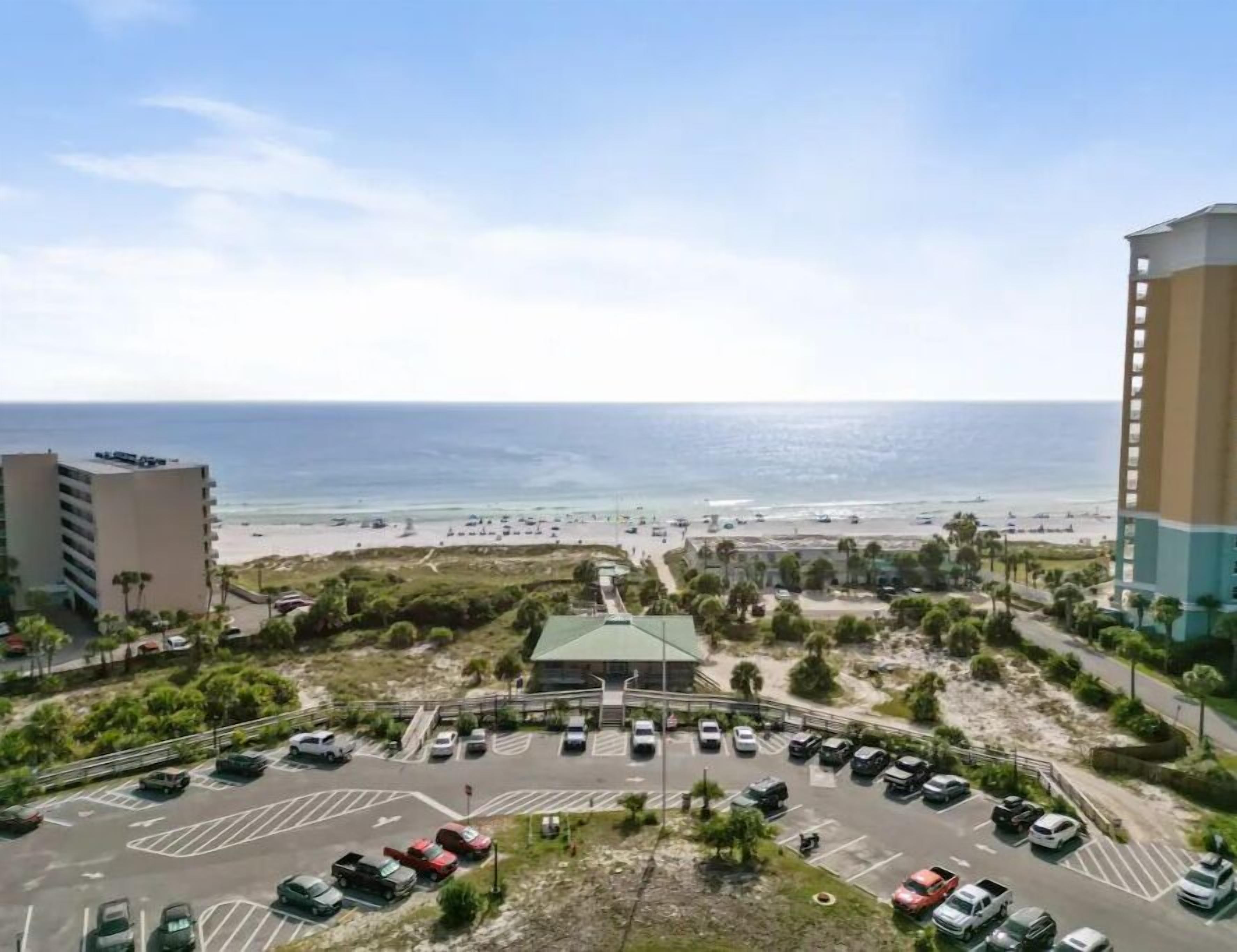 A view of a parking lot with several cars, a building with a green roof, and a beach in the background.