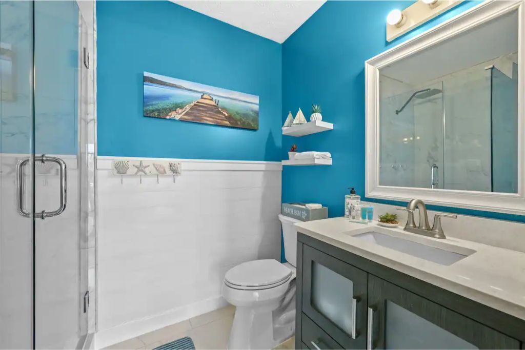 A bathroom with a blue wall, a glass shower enclosure, a white toilet, a sink with a mirror, and a shelf with toiletries.