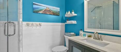 A bathroom with a blue wall, a glass shower enclosure, a white toilet, a sink with a mirror, and a shelf with toiletries.