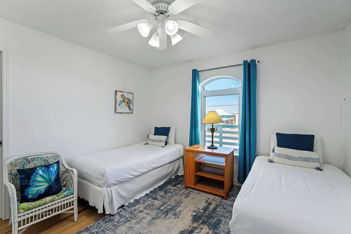 A room with two single beds, a chair with a butterfly cushion, a small table with a lamp, and a window with curtains.