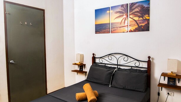 Un dormitorio con una cama, una puerta, una mesita de noche con una lámpara y una pared con una pintura de atardecer.