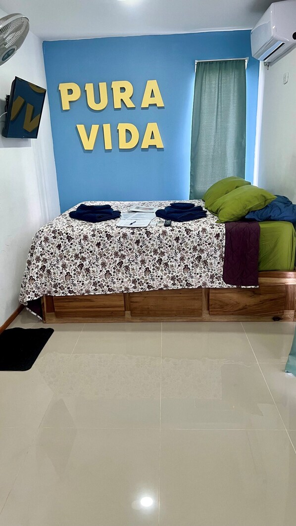 A bedroom with a bed, a blue wall with 'PURA VIDA' text, a window with curtains, and an air conditioning unit. - Kingston Casitas Private 3rd Flr, #6 with AC, Free Bikes… (Puerto Viejo de talamanca)