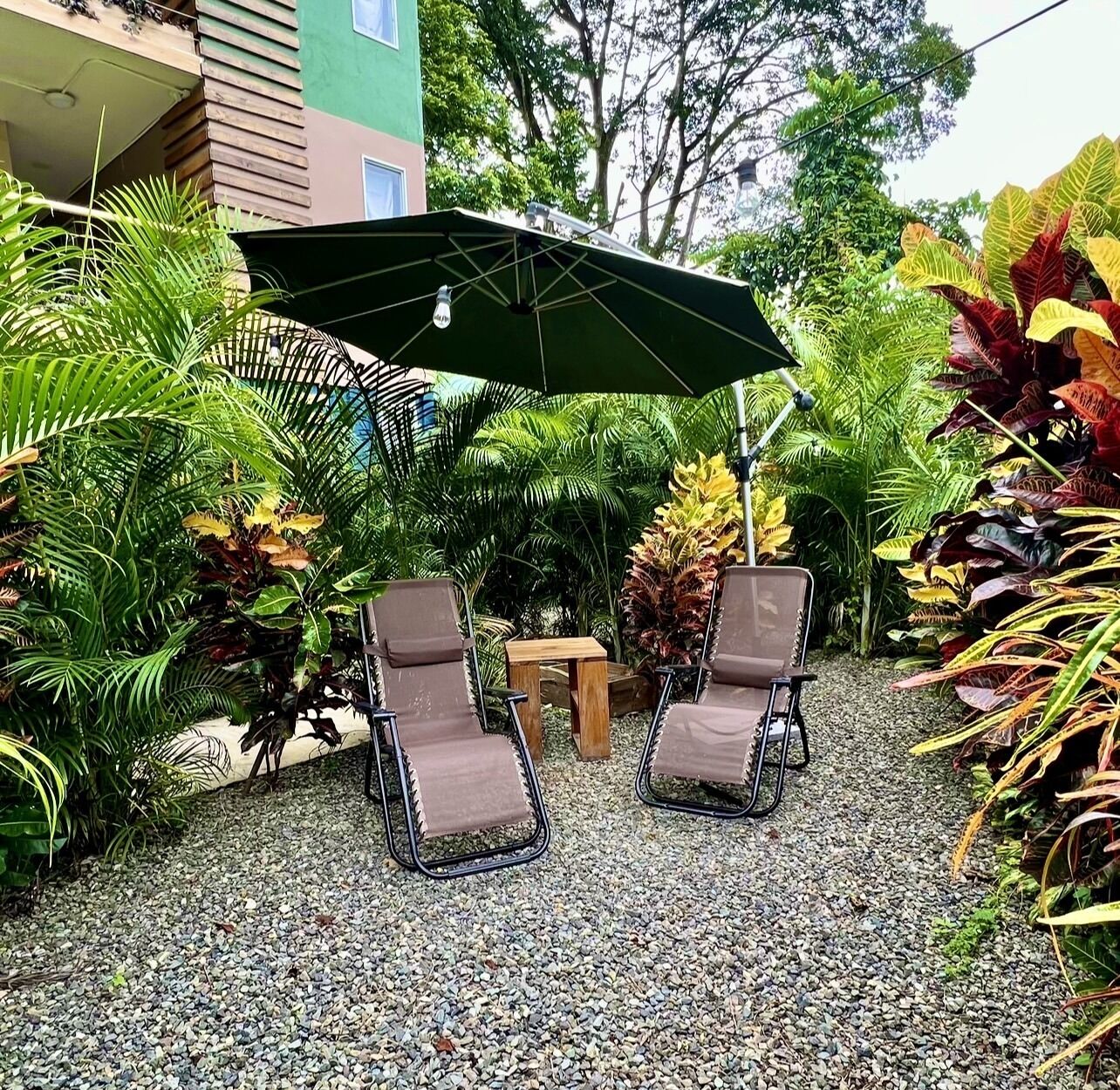 An outdoor seating area with two lounge chairs and a table under a green umbrella, surrounded by lush tropical plants.