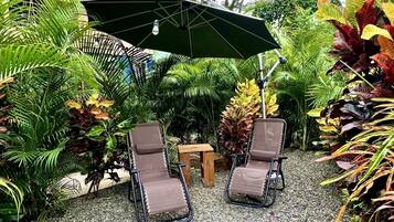 An outdoor seating area with two lounge chairs and a table under a green umbrella, surrounded by lush tropical plants.