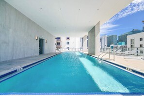 An indoor swimming pool with clear blue water, surrounded by a modern building with balconies and a view of high-rise buildings.