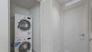 A compact laundry room with a front-loading washer and dryer, a white door, and a tiled floor. - Designer 1BR | 24th Floor Gym | Brickell Core (Miami)
