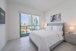 A modern hotel room with a large bed, a nightstand, a painting, and a city view through a large window.
