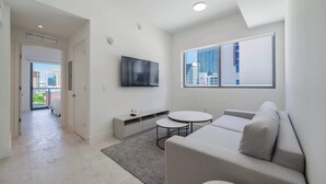 A modern living room with a grey sofa, a flat-screen TV, a small round table, and a view of city buildings through a window. - Designer 1BR | 24th Floor Gym | Brickell Core (Miami)