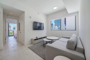 A modern living room with a grey sofa, a flat-screen TV, a small round table, and a view of city buildings through a window.