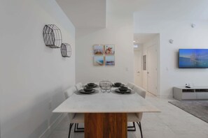 A modern dining area with a white table and chairs, a wooden accent on the table, and a TV mounted on the wall.