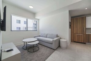 Ein modernes Wohnzimmer mit grauem Sofa, einem kleinen runden Tisch und einem weißen Couchtisch. Ein großes Fenster bietet einen Ausblick nach draußen.