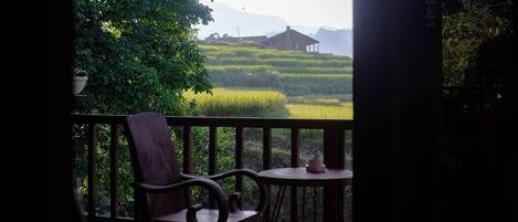 A balcony with a wooden chair and table, overlooking a green field and distant mountains.