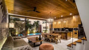 Una cocina moderna con armarios de madera y una isla central, un área de comedor con una mesa grande y sillas, y una zona de estar acogedora con sofá y reposapiés.