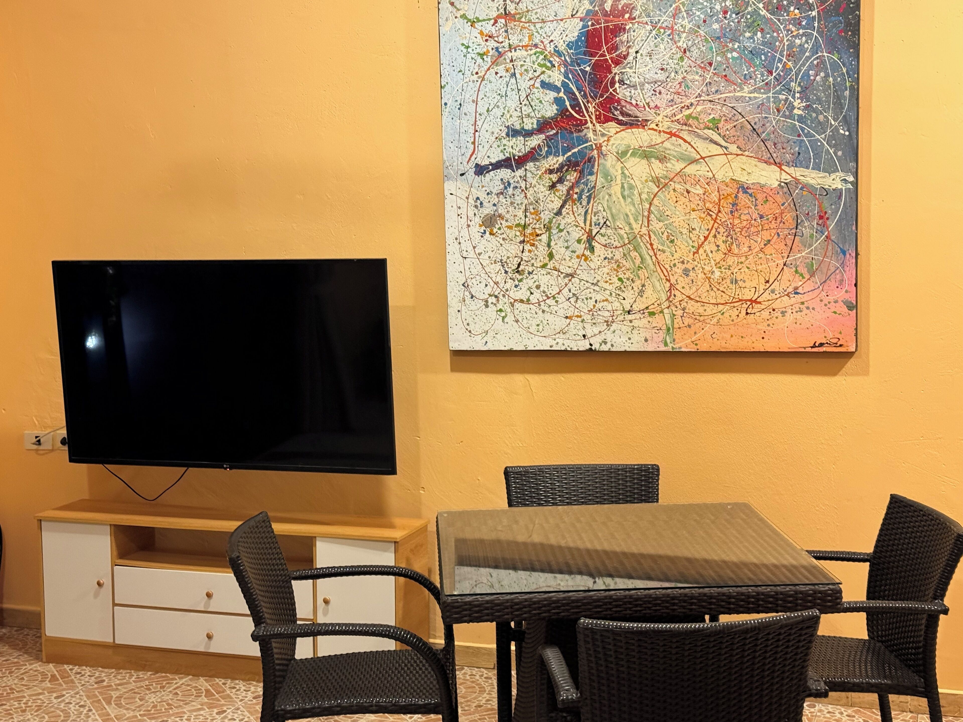 Quarto com televisão de tela plana presa a um armário de madeira, uma mesa de jantar com quatro cadeiras de vime preto e um quadro abstrato colorido na parede.