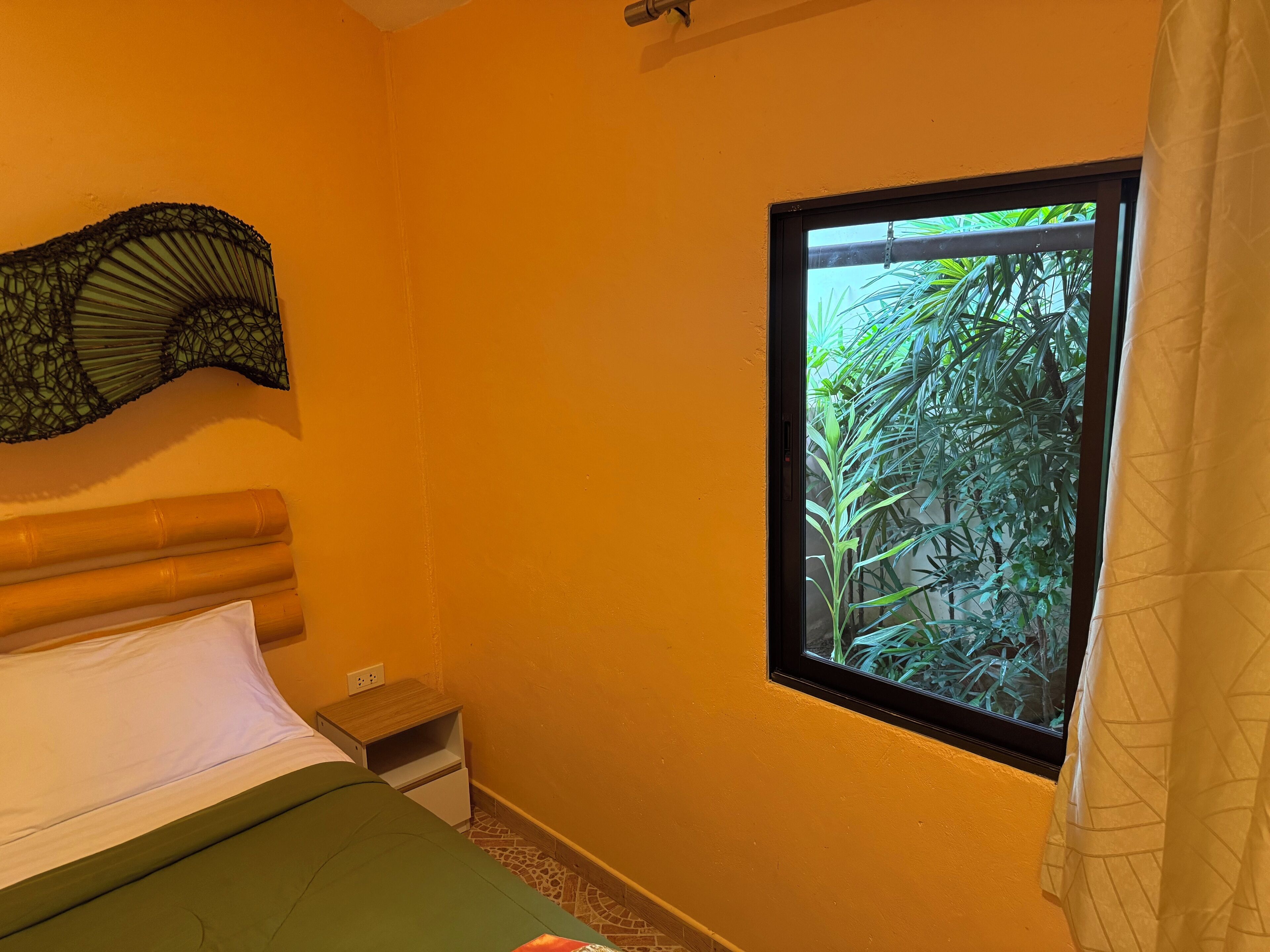 A bedroom with a bed, a nightstand, a window with a view of greenery, and a wall-mounted decorative item.