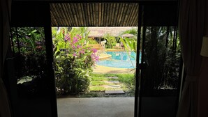 Une chambre avec vue sur une piscine, entourée d’une végétation luxuriante et surmontée d’une structure à toit de chaume.