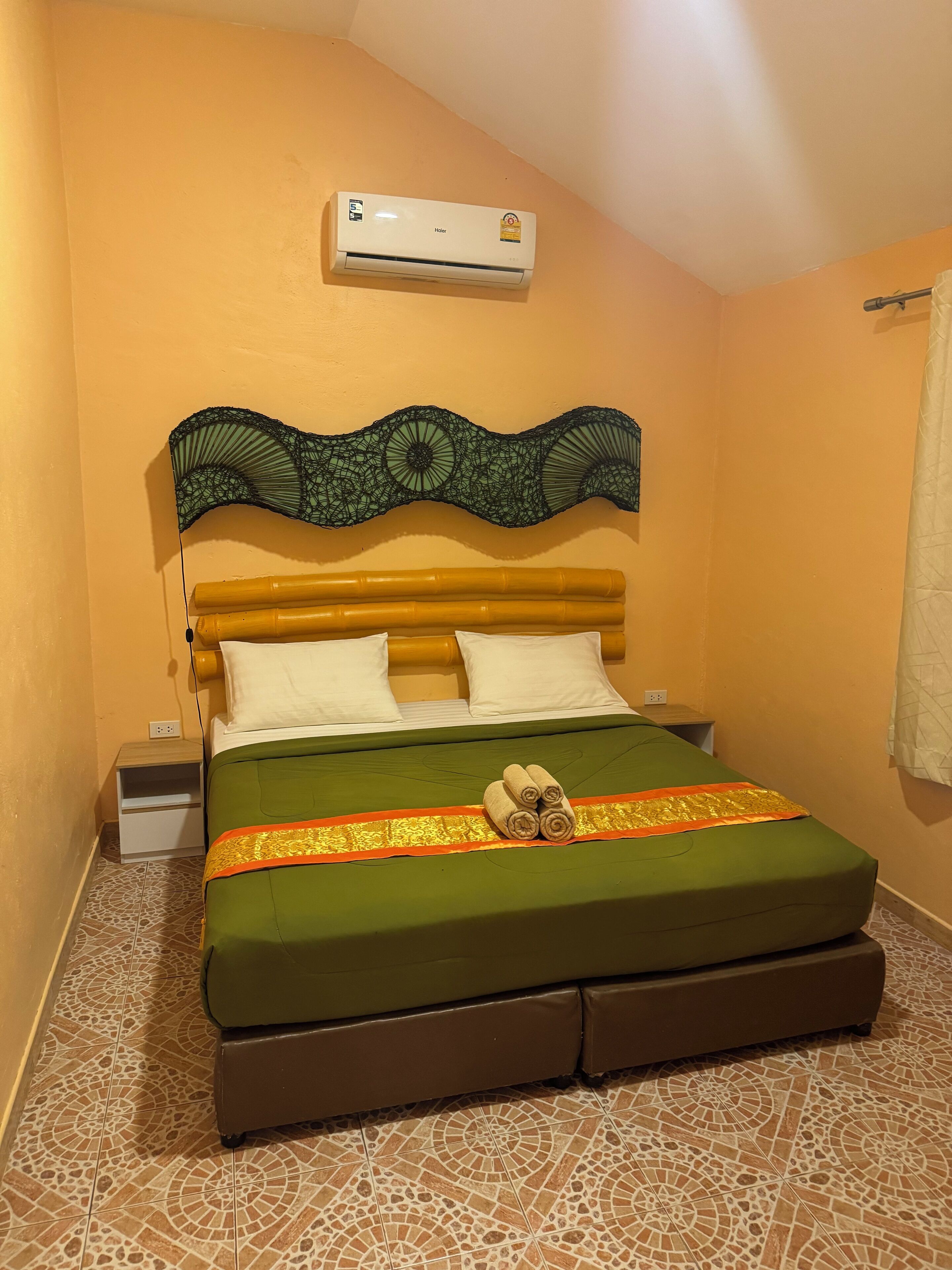 A hotel room with a bed, green bedspread, two white pillows, a decorative headboard, an air conditioner, a nightstand, and a patterned floor.