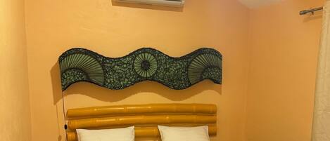 A hotel room with a bed, green bedspread, two white pillows, a decorative headboard, an air conditioner, a nightstand, and a patterned floor.