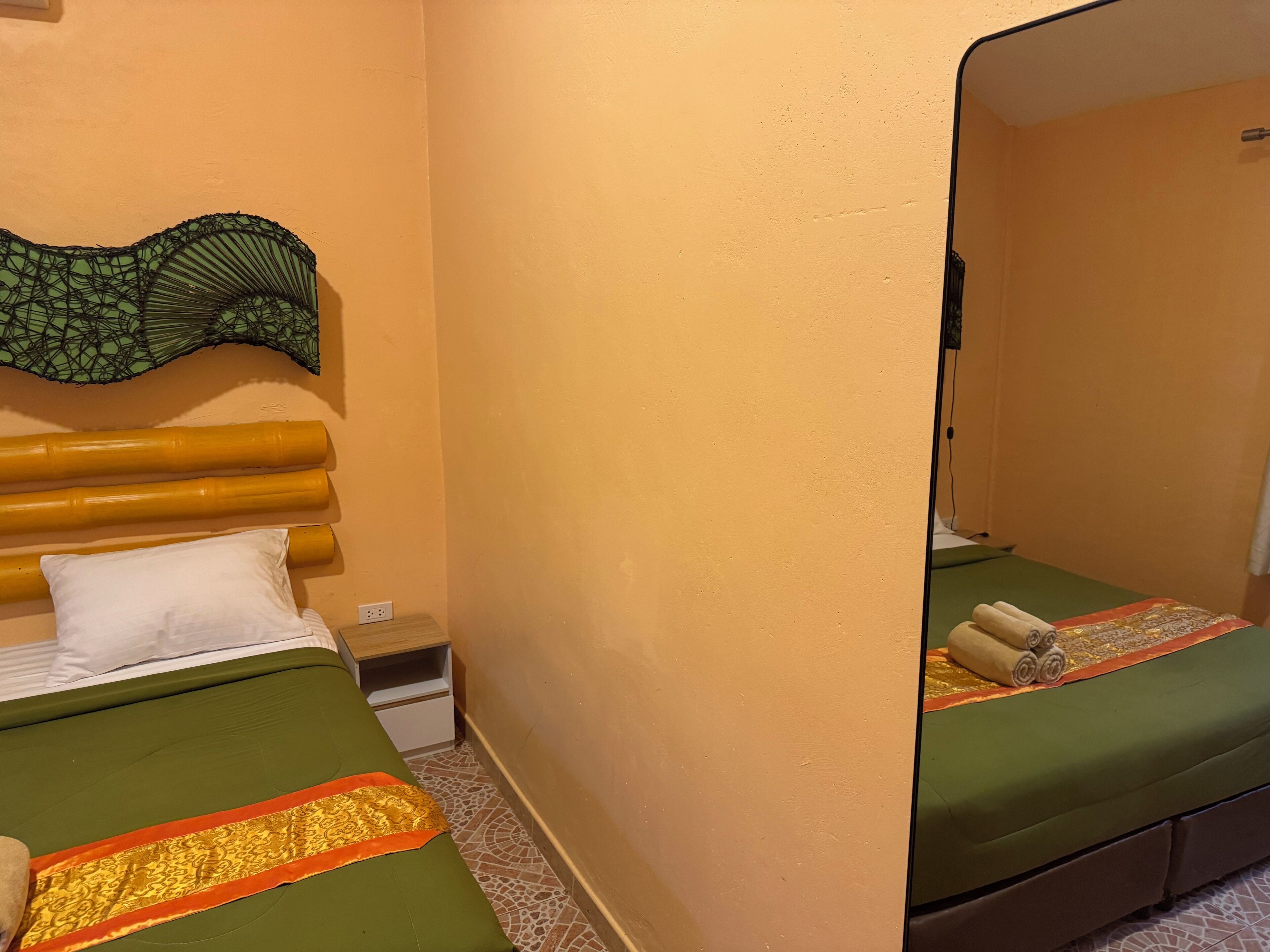 A room with two beds, a green bedspread, a white pillow, a wooden headboard, a small nightstand, and a wall-mounted mirror.