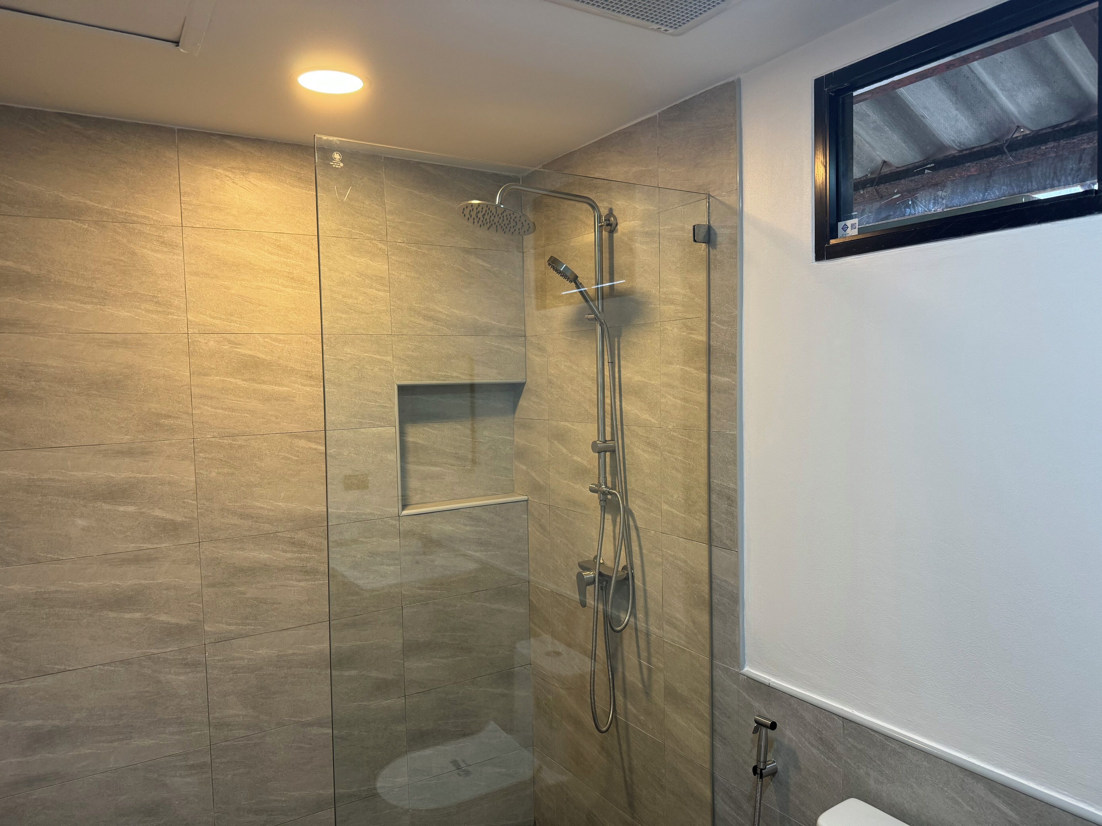 Un baño moderno con ducha envuelta en vidrio, estante empotrado y ventana sobre el lavabo.