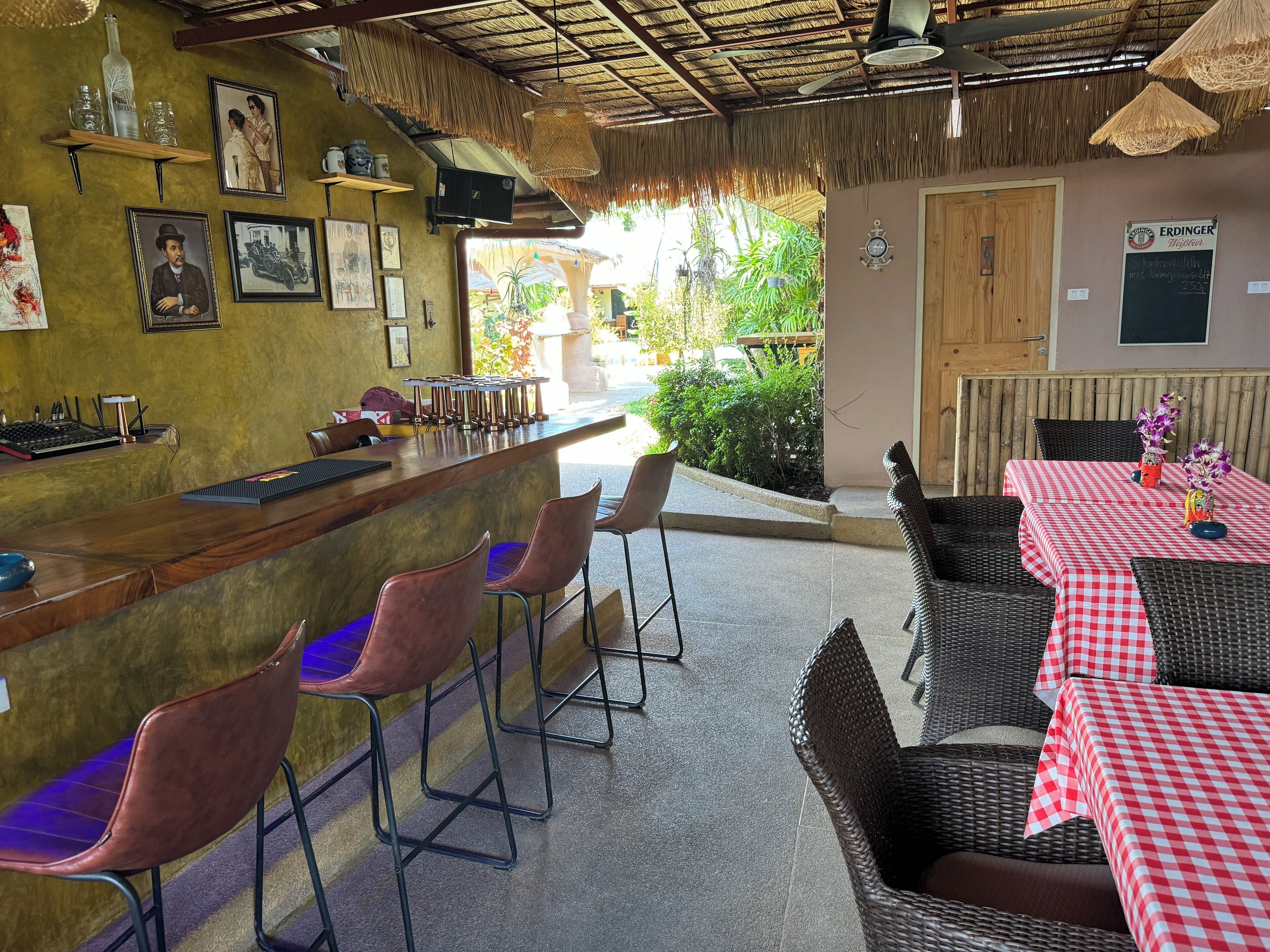 Un área de bar con sillas y mesas de mimbre, cuadros en la pared y un techo de paja.