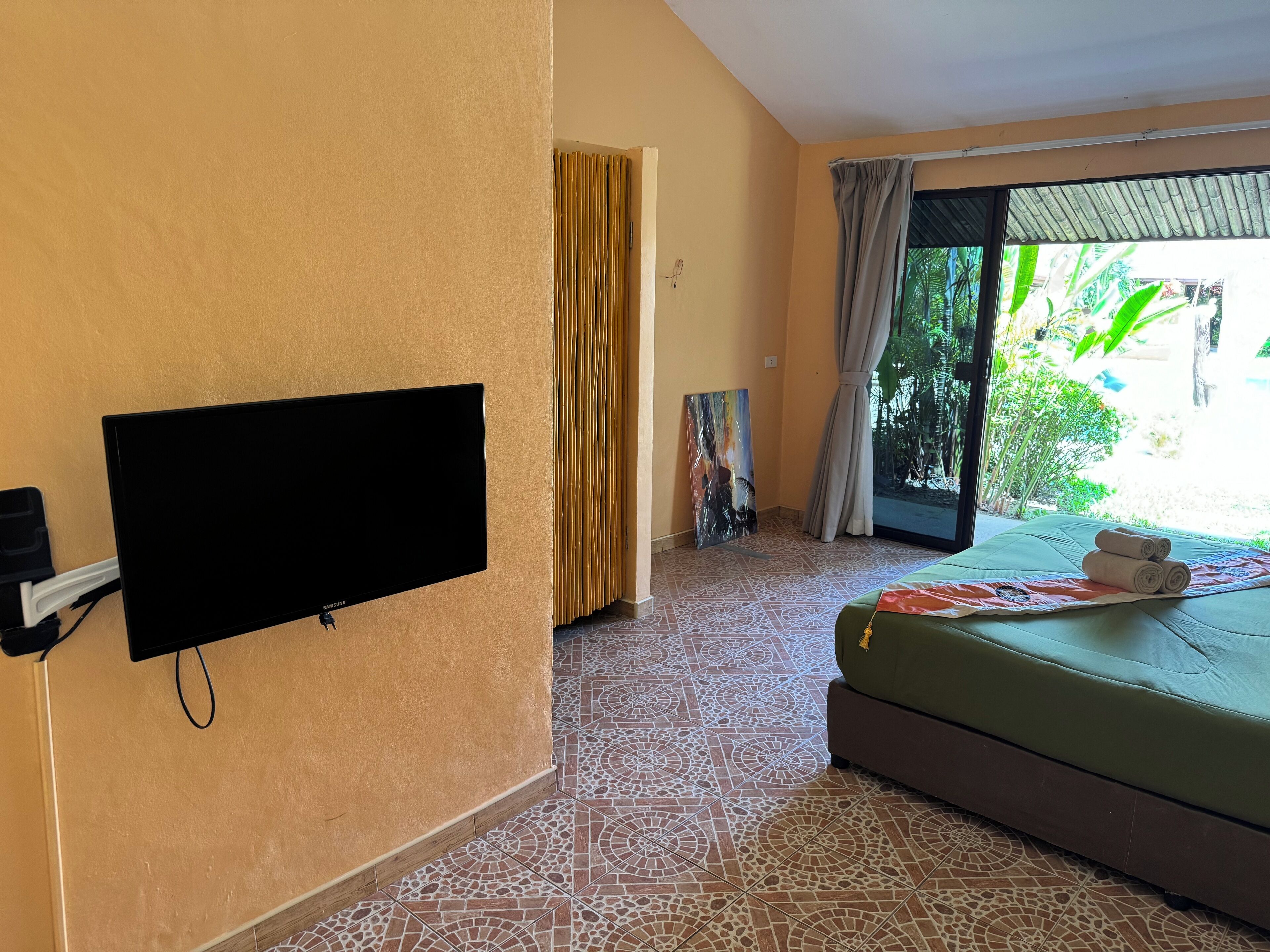 Habitación con televisión de pantalla plana montada en la pared, una cama con colchón verde y una toalla enrollada, un cuadro en la pared y vistas a la vegetación por la ventana.
