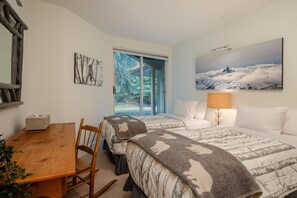 Ein Schlafzimmer mit zwei Betten, einem Holztisch, einem Stuhl, einer Lampe und einem Gemälde einer Berglandschaft.