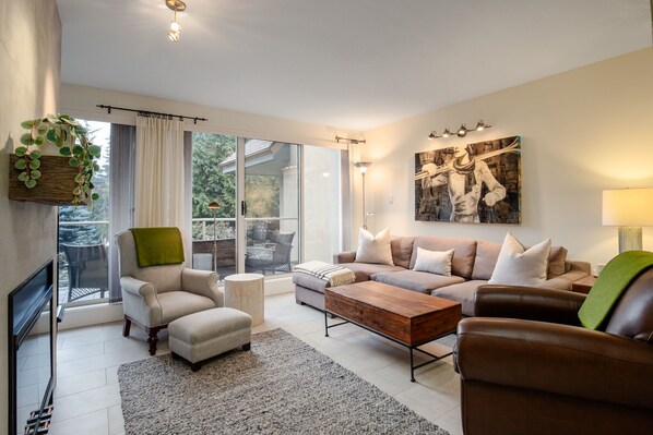Ein modernes Wohnzimmer mit einer Sofagarnitur, einem Couchtisch und einem Beistelltisch. Ein großes Fenster mit Vorhängen, eine Pflanze und ein Gemälde an der Wand sind zu sehen.