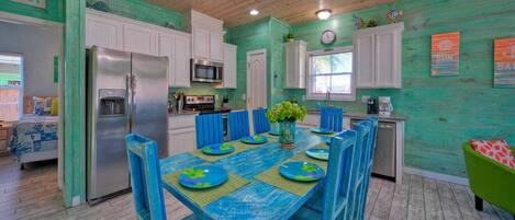 Une cuisine aux murs de couleur turquoise, avec des meubles blancs et un coin repas doté de chaises et d’assiettes bleues.