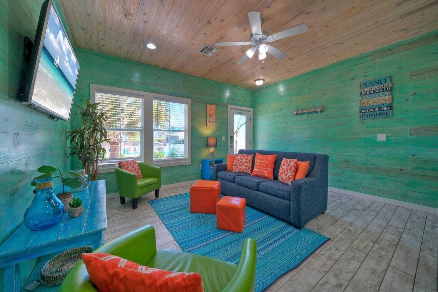 A room with a wooden ceiling, a blue sofa with orange cushions, a green armchair, and a blue coffee table. There is a TV on the wall and a window with blinds.