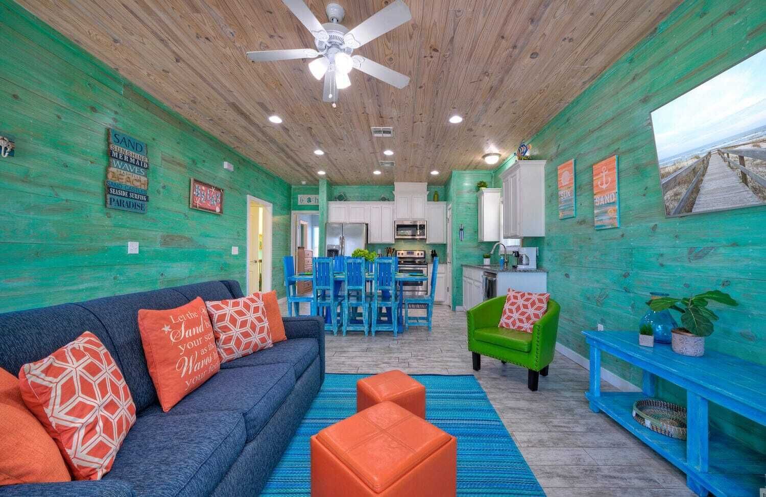 A vibrant room with a blue sofa, orange ottomans, and a green armchair. A ceiling fan and wooden ceiling are visible.