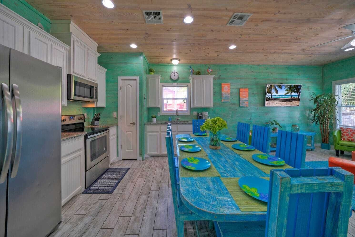 A kitchen with white cabinets, a stainless steel refrigerator, a microwave, and a stove. There is a dining area with blue chairs and a table set with plates and a vase of flowers.