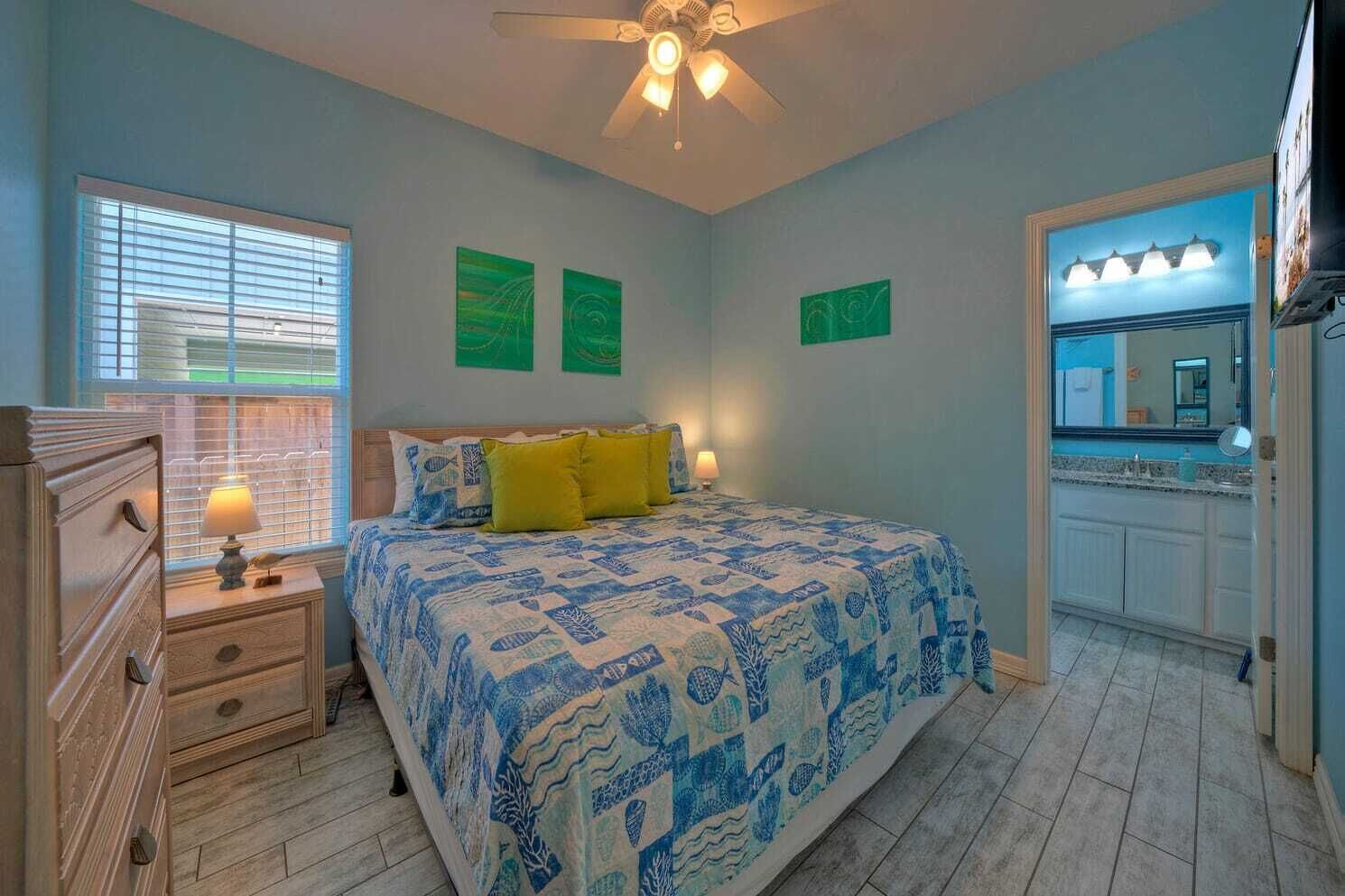 Une chambre à coucher comprenant un lit, une table de chevet, une commode, un ventilateur de plafond et une salle de bain visible à travers une porte ouverte.