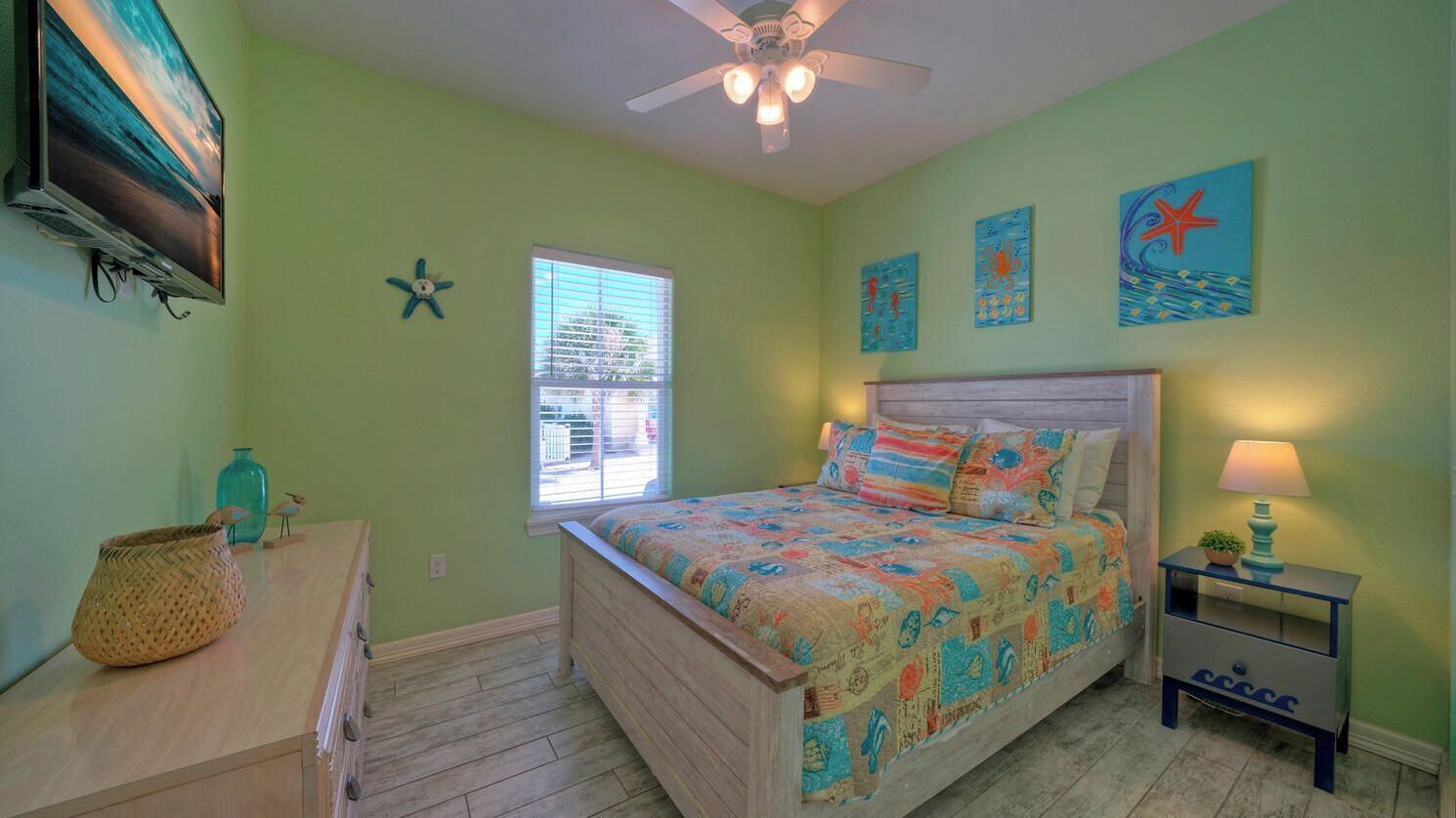 A bedroom with a bed, a dresser, a nightstand, a ceiling fan, and artwork on the walls.