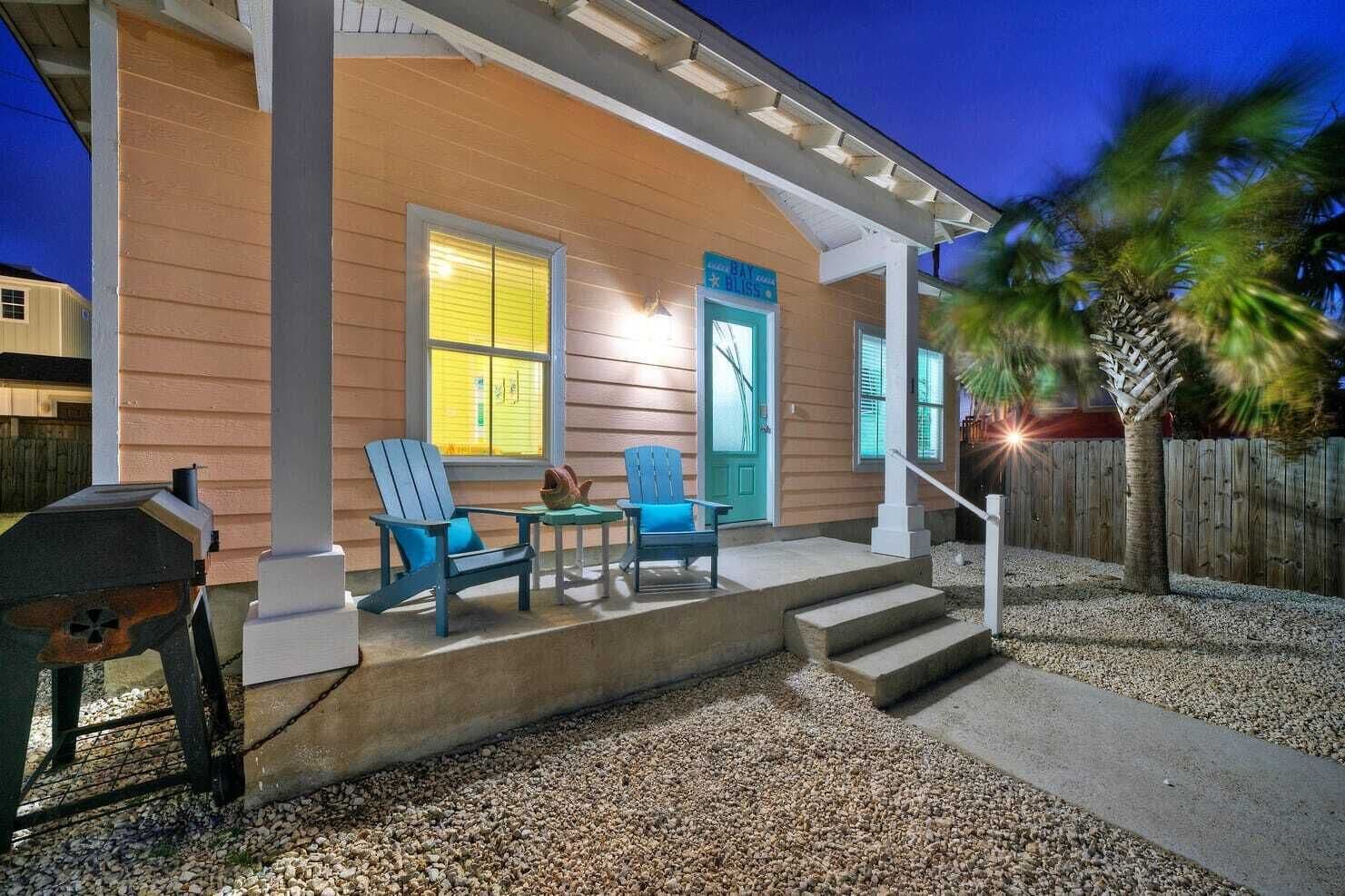 A house with a porch, two blue Adirondack chairs, a grill, and a wooden fence.