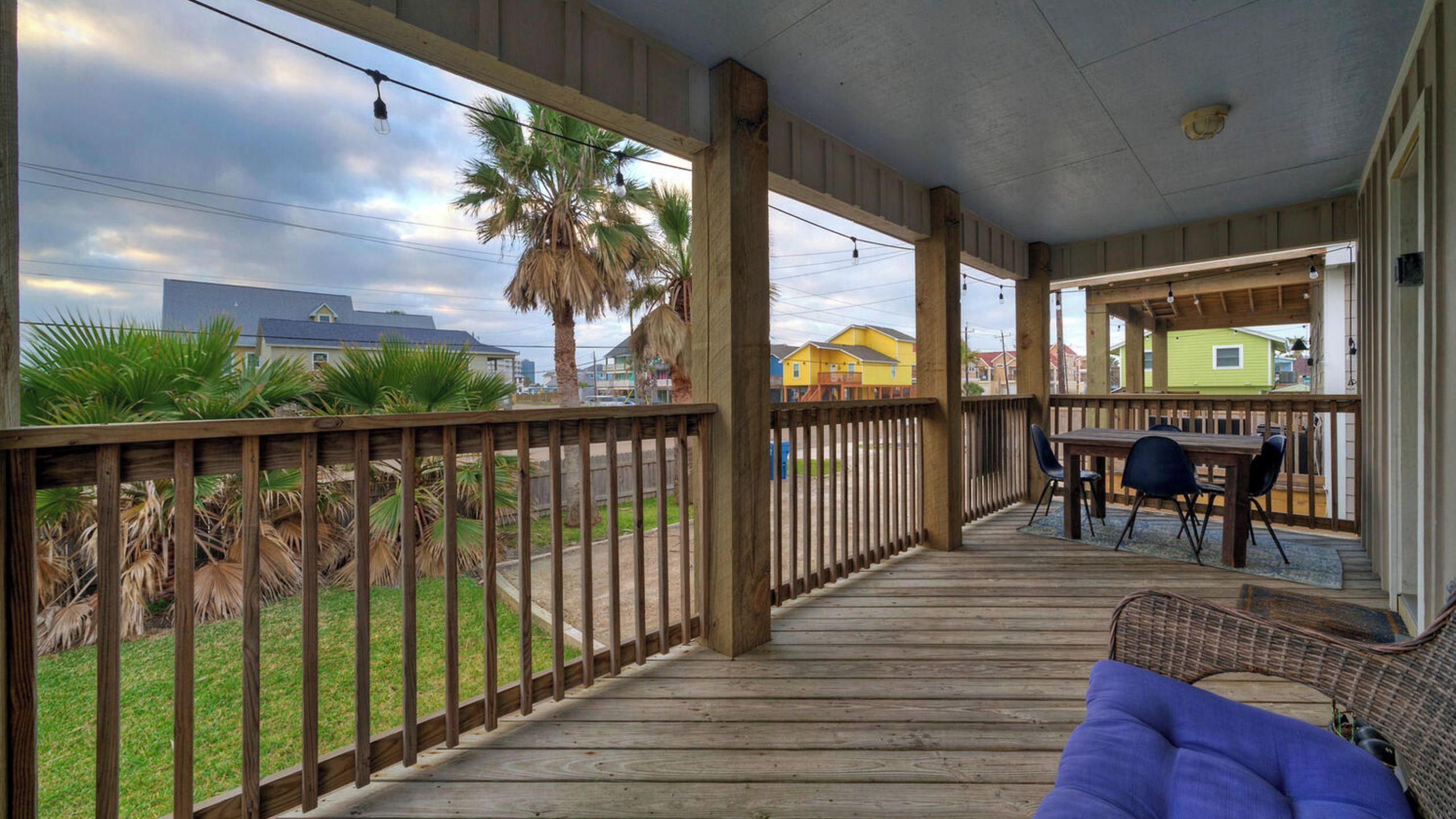 A covered porch with a wooden deck, a dining area with chairs, and a view of houses and palm trees.