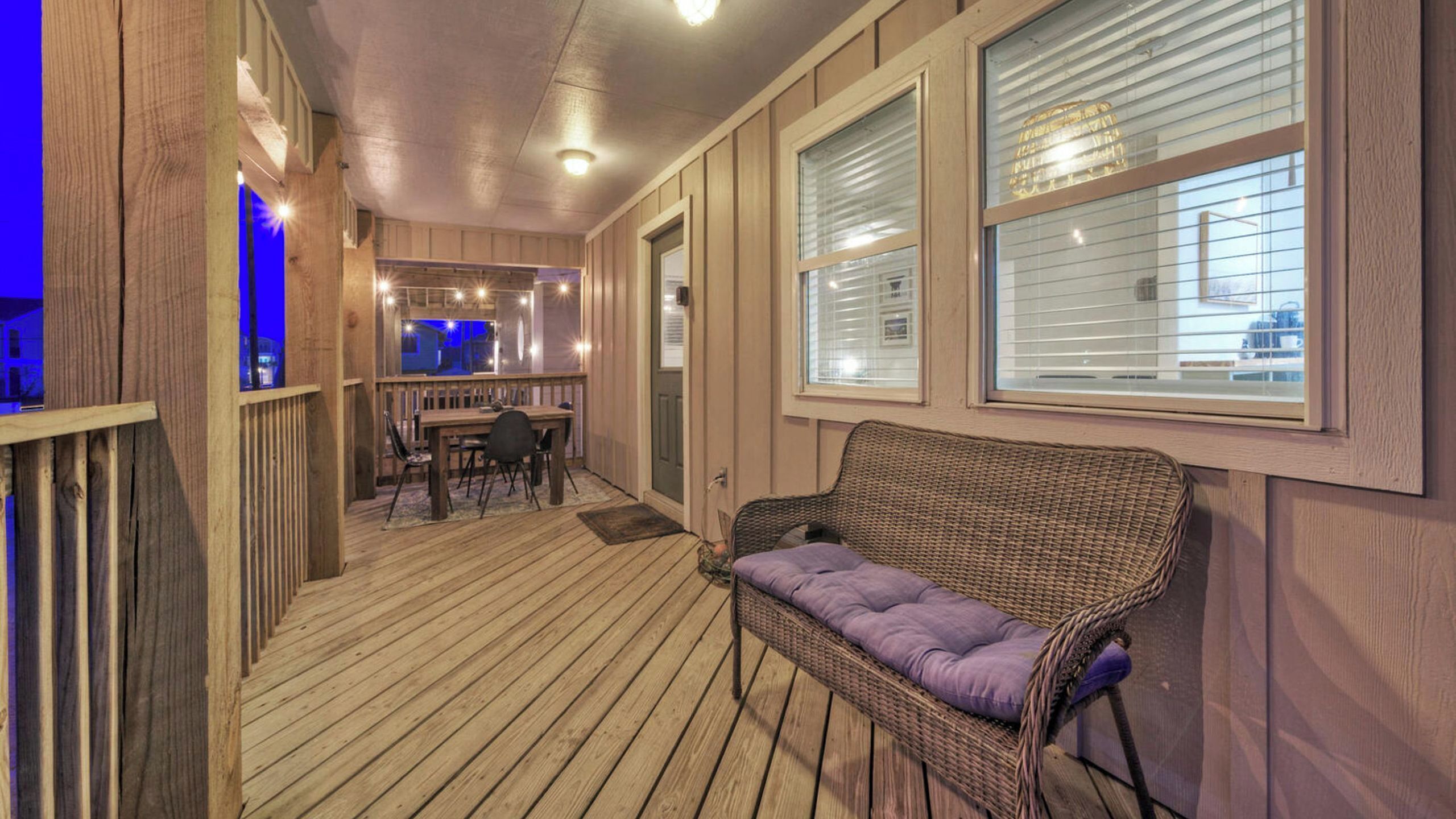 A covered outdoor area with a wooden deck, a wicker sofa, a dining table with chairs, and a bar area with lights.