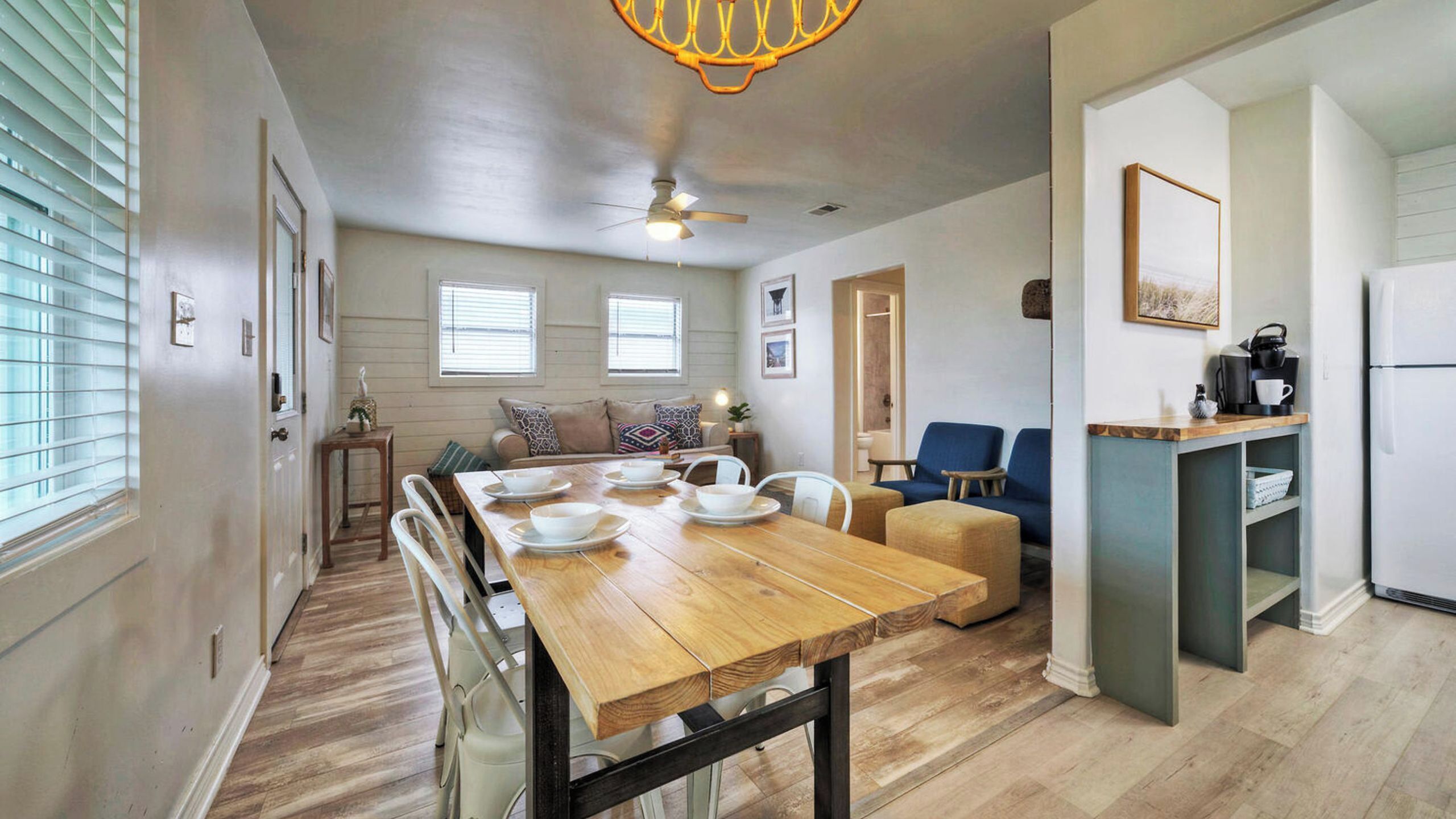 A dining area with a wooden table and chairs, a ceiling fan, and a kitchen area with a refrigerator and coffee maker.