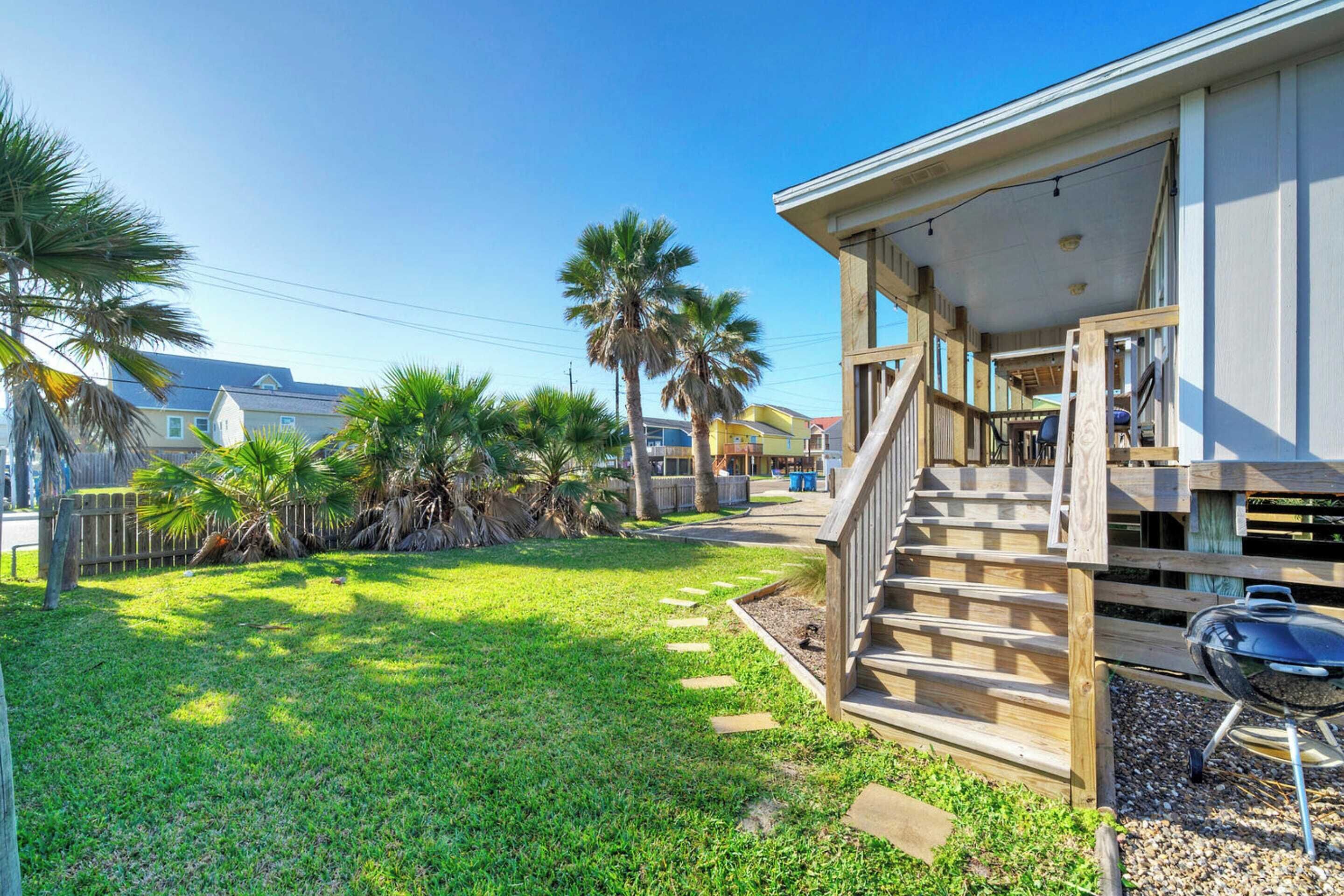 A residential property with a wooden staircase leading to a covered porch, a barbecue grill, and a well-maintained lawn with palm trees.