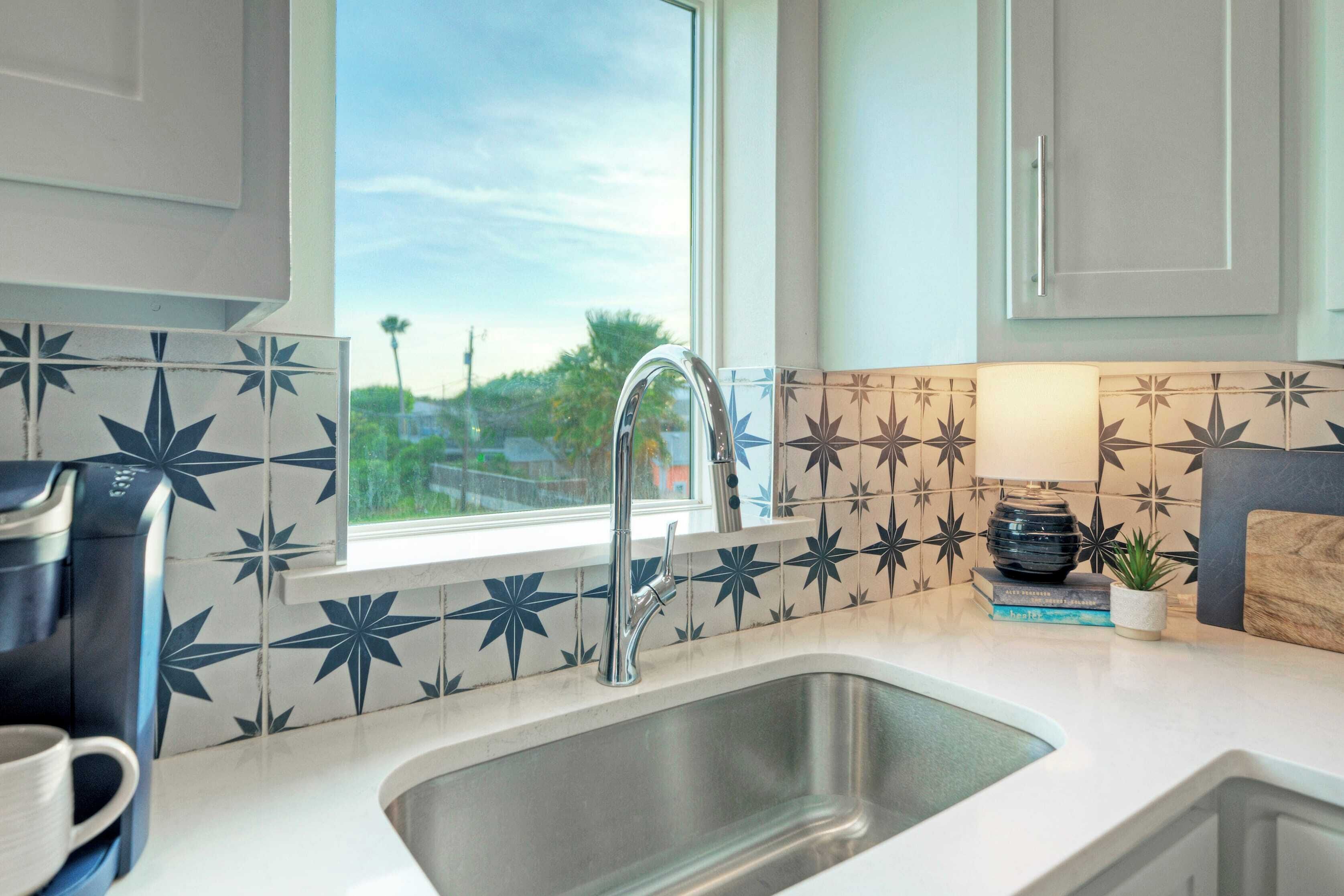 A kitchen with a stainless steel sink, a coffee maker, and a window with a view of greenery.