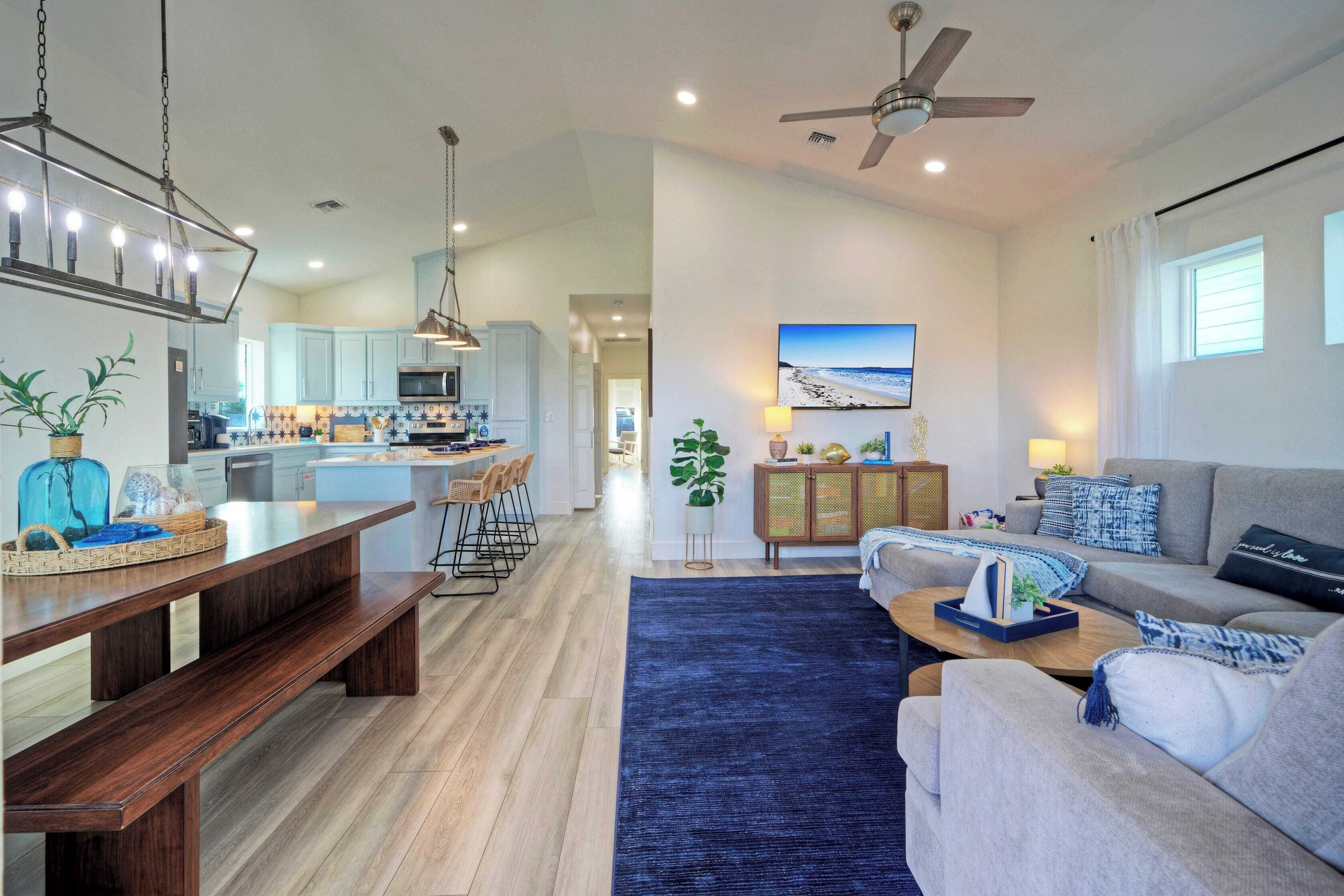 A spacious living area with a large sofa, a coffee table, and a dining table with benches. There is a kitchen in the background with light blue cabinets and a ceiling fan.