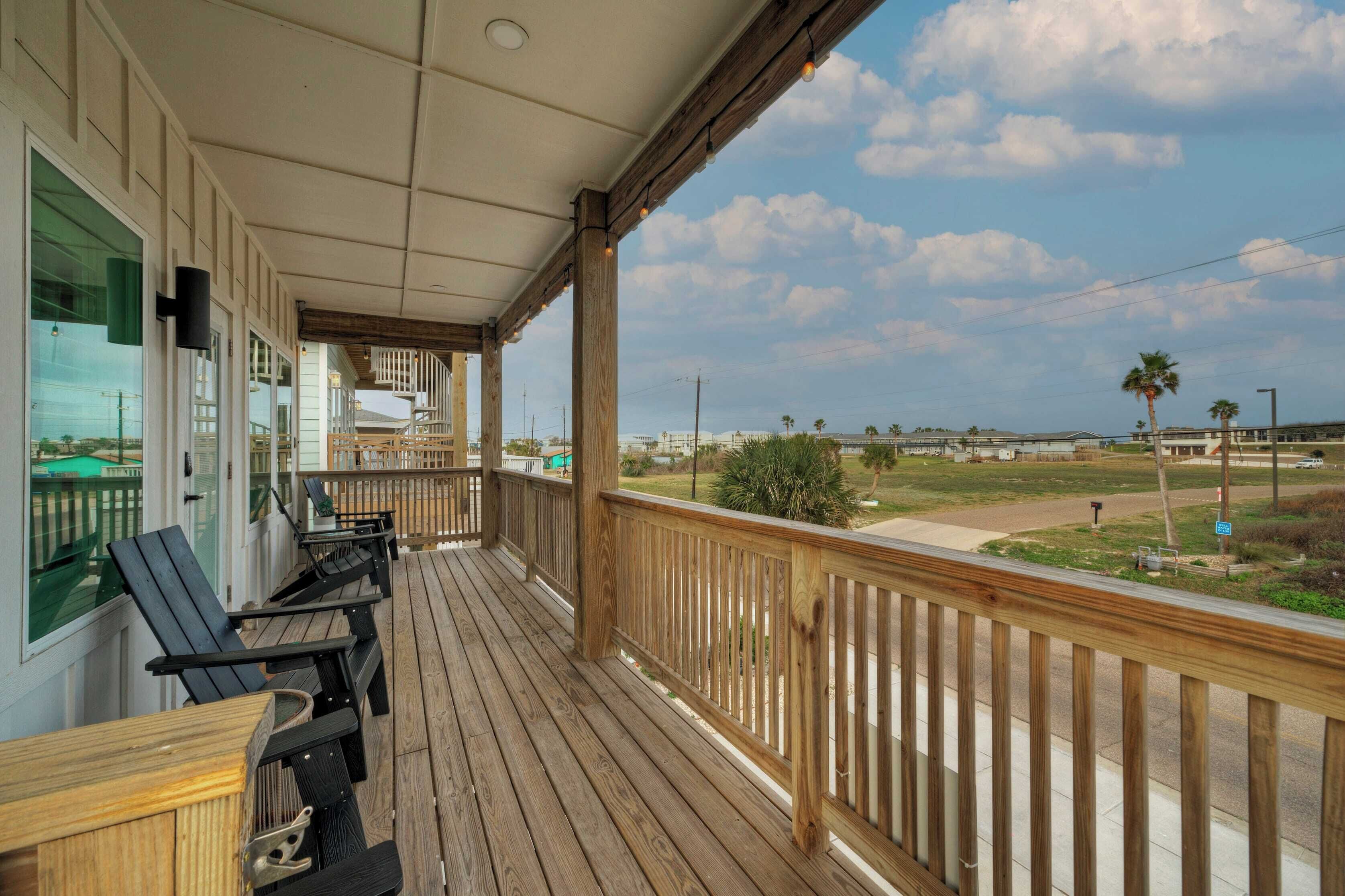A wooden deck with outdoor seating, overlooking a grassy area with palm trees and a distant view of buildings.