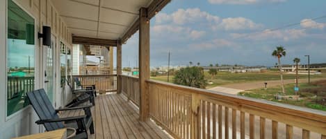 A wooden deck with outdoor seating, overlooking a grassy area with palm trees and a distant view of buildings.