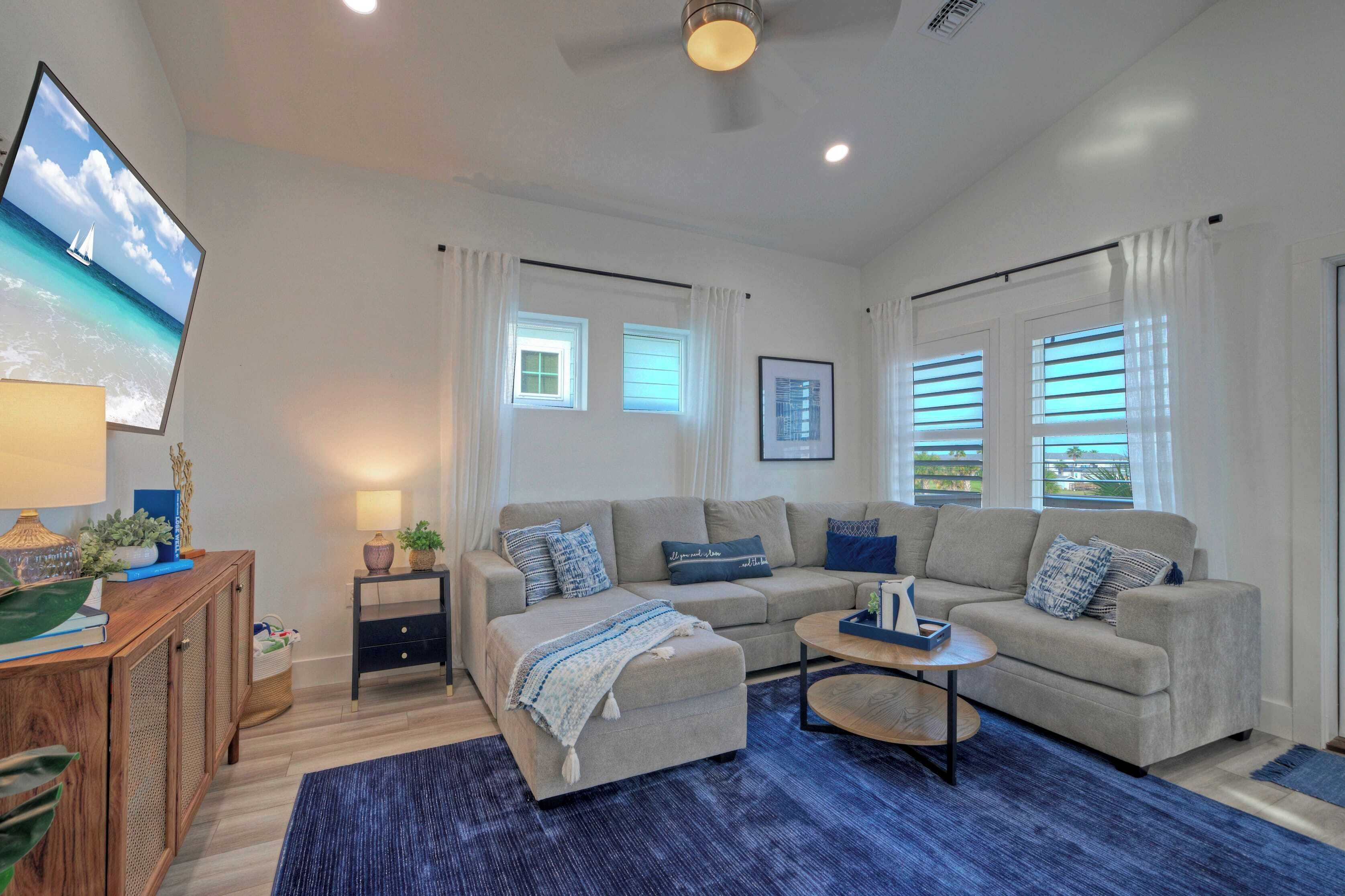 A modern living room with a sofa, ottoman, and coffee table, a TV mounted on the wall, and a window with blinds.