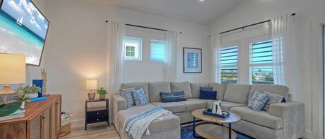 A modern living room with a sofa, ottoman, and coffee table, a TV mounted on the wall, and a window with blinds.