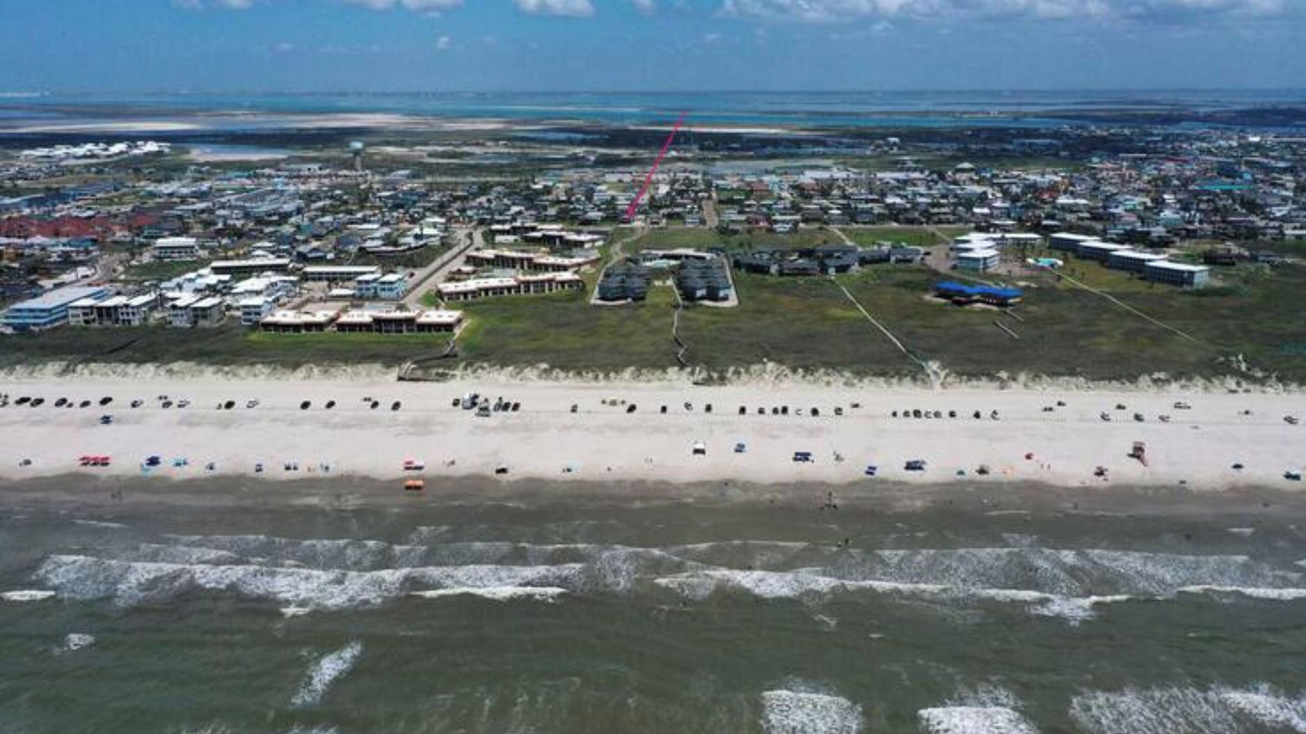 An aerial view of a coastal area with a beach, buildings, and a road.