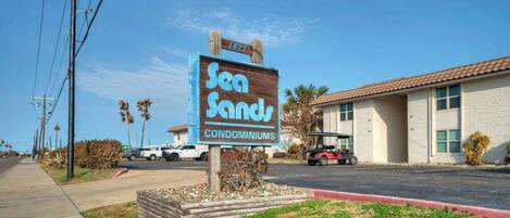 A sign for Sea Sands Condominiums with a clear view of the building and a parking area.